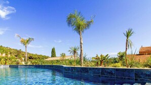A swimming pool with clear blue water, surrounded by palm trees and greenery, with a view of a hillside and a building with a red roof in the background. - Bright apartment with large terrace and pool, Le Lavandou (Le Lavandou)
