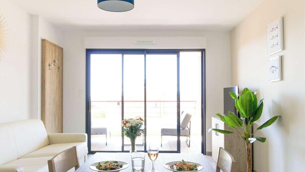 A dining area with a white table set for two, a sofa, and a large window with a view of the outdoors. - Bright apartment with large terrace and pool, Le Lavandou (Le Lavandou)