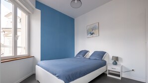 A bedroom with a bed, a nightstand, a lamp, a painting, and a window. - Apartment in Le Mans Near City Center (Le Mans)