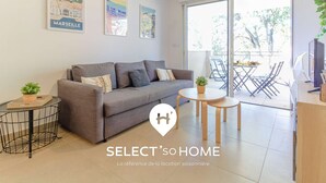 A modern living room with a grey sofa, a small round table, and a TV. There are framed pictures on the wall and a balcony with outdoor furniture.