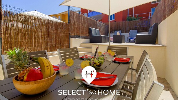 Un área de comedor al aire libre con una mesa dispuesta para dos personas, que incluye un cuenco de madera con frutas, un vaso de agua y una servilleta roja.