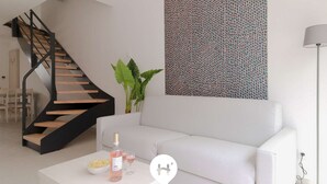 Una sala moderna con un sofá blanco, una mesa de centro con una botella y vasos, una escalera con escalones de madera y una pared con un diseño.