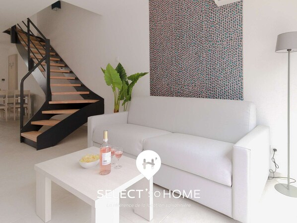 Una sala moderna con un sofá blanco, una mesa de centro con una botella y vasos, una escalera con escalones de madera y una pared con un diseño.