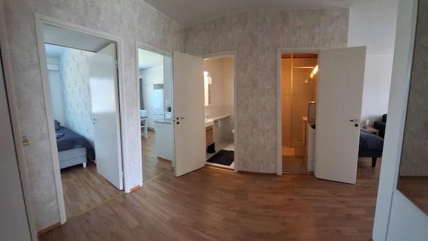 Een ruime kamer met houten vloeren, een zwart tapijt en een zichtbare badkamer met douche.