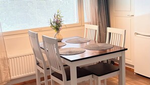 Een eetgedeelte met een tafel voor vier personen, witte stoelen, een vaas met bloemen en een raam met jaloezieën.