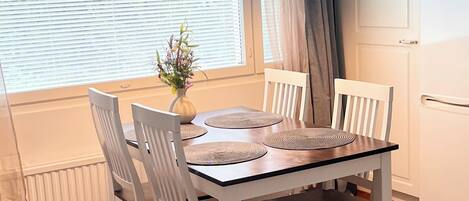 Un comedor con una mesa dispuesta para cuatro, sillas blancas, un florero con flores y una ventana con persianas.