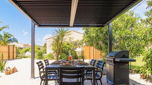 Une terrasse extérieure dotée d’un espace salon couvert, d’un barbecue et d’une table dressée pour un repas.