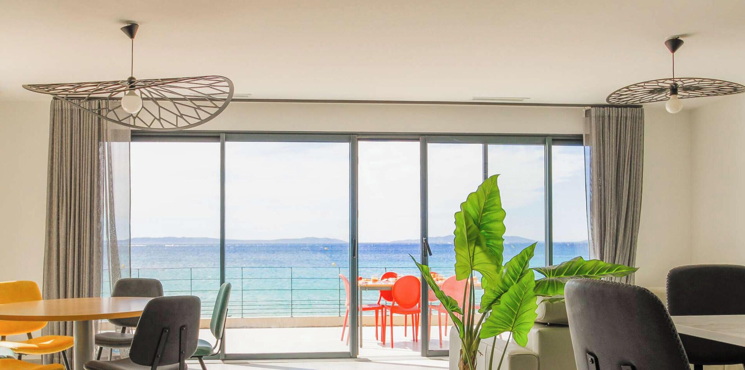 A modern room with a large sliding glass door leading to a balcony with a view of the sea. There are several chairs and a table inside the room.