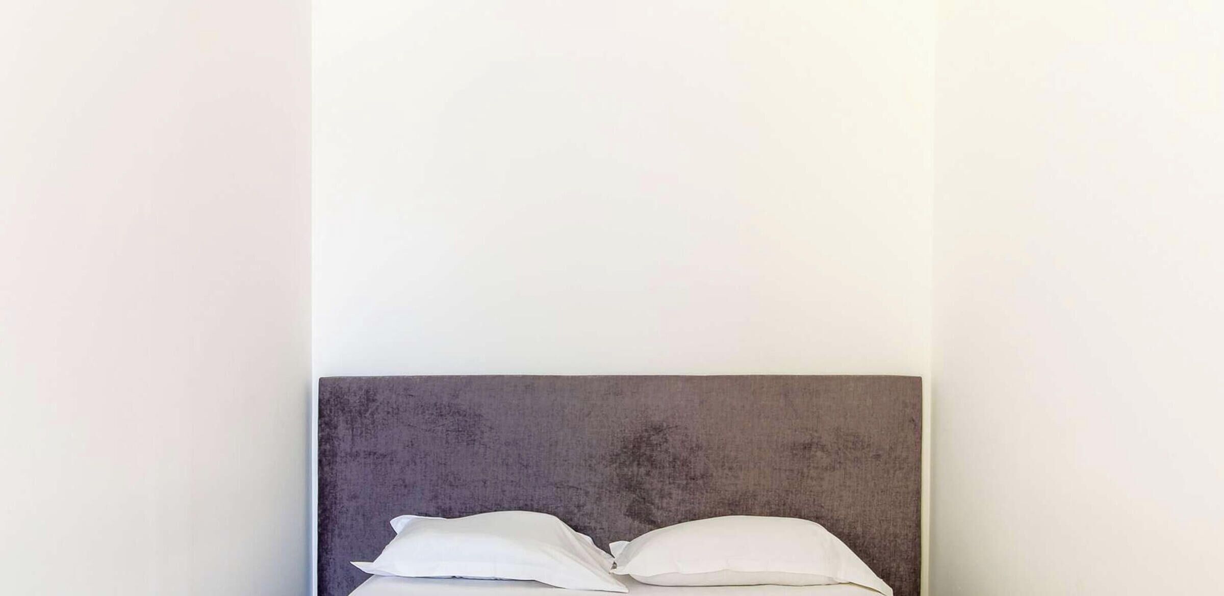 Ein Bett mit grauem Kopfteil und weißen Kissen vor einer schlichten Wand.