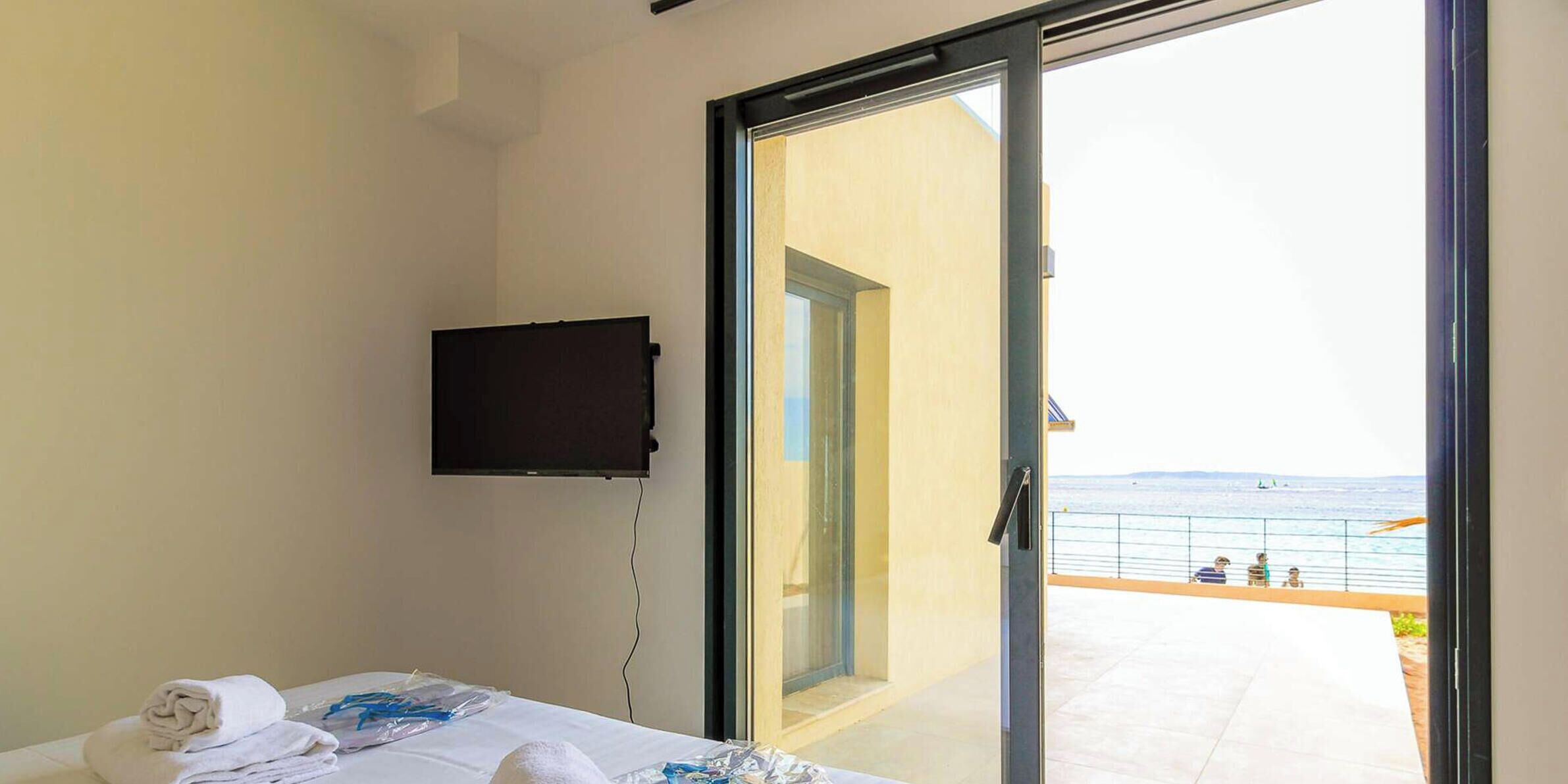 A hotel room with a bed, a flat-screen TV mounted on the wall, and a sliding glass door leading to a balcony with a view of the sea.