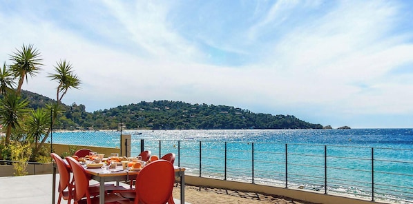 Une table entourée de chaises et recouverte d’une nappe, installée sur une terrasse donnant sur une plage, avec une montagne en arrière-plan.