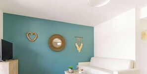 Ein Wohnzimmer mit türkisfarbenen Wänden, einem weißen Sofa, einem Holz-TV-Schrank und Wanddekorationen, darunter ein herzförmiger Spiegel und ein Wandbehang aus Makramee.