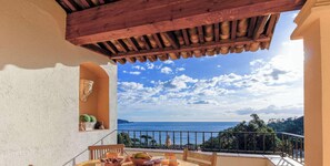Un balcón con vista al mar, una mesa con frutas y un cuenco, y una planta en maceta.