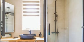 Ein Badezimmer mit großem Spiegel, einem runden Waschbecken, einer Duschbrause und einem Fenster mit Jalousien.