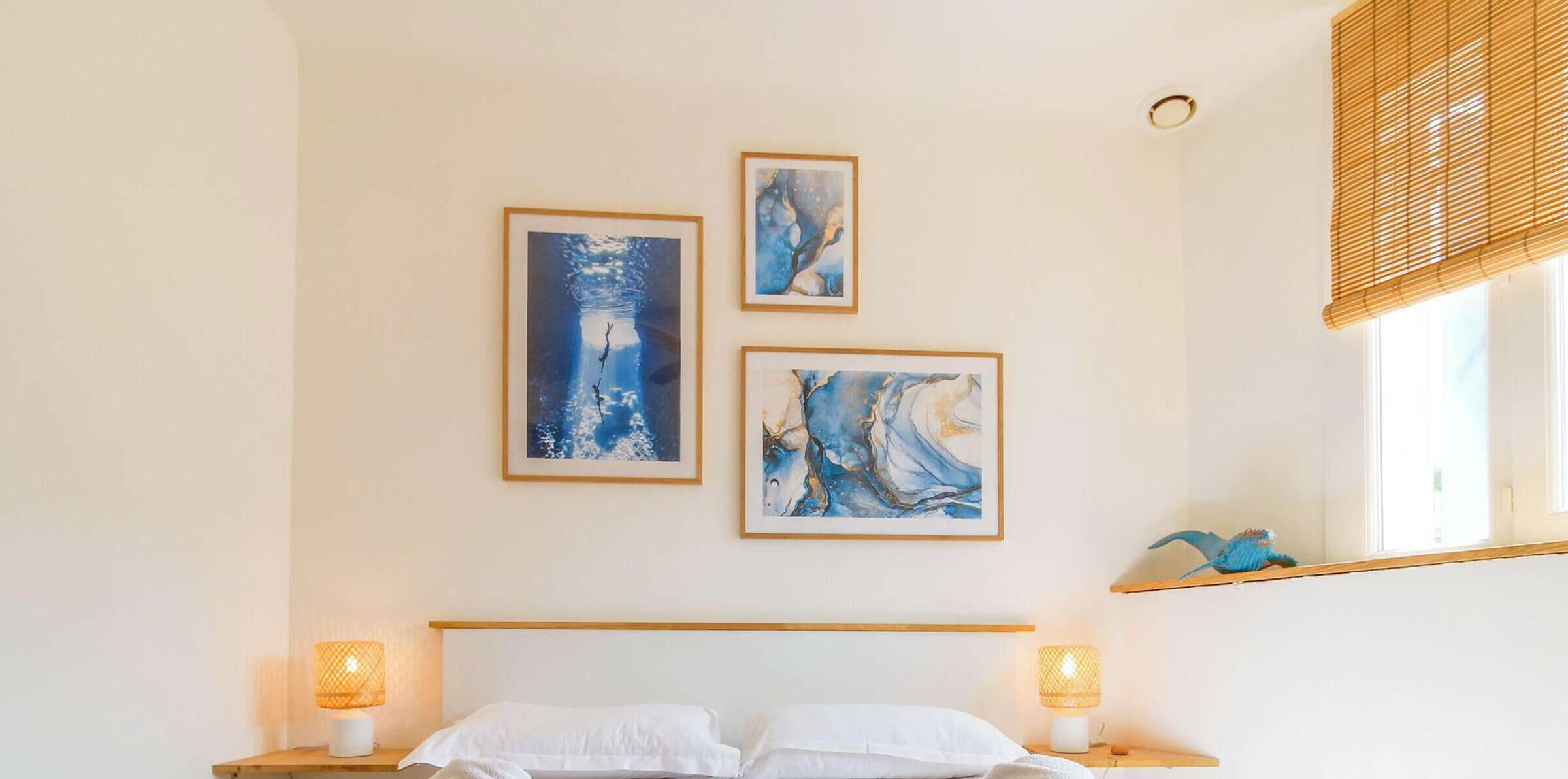 A bedroom with a bed, two bedside lamps, and three framed artworks on the wall.