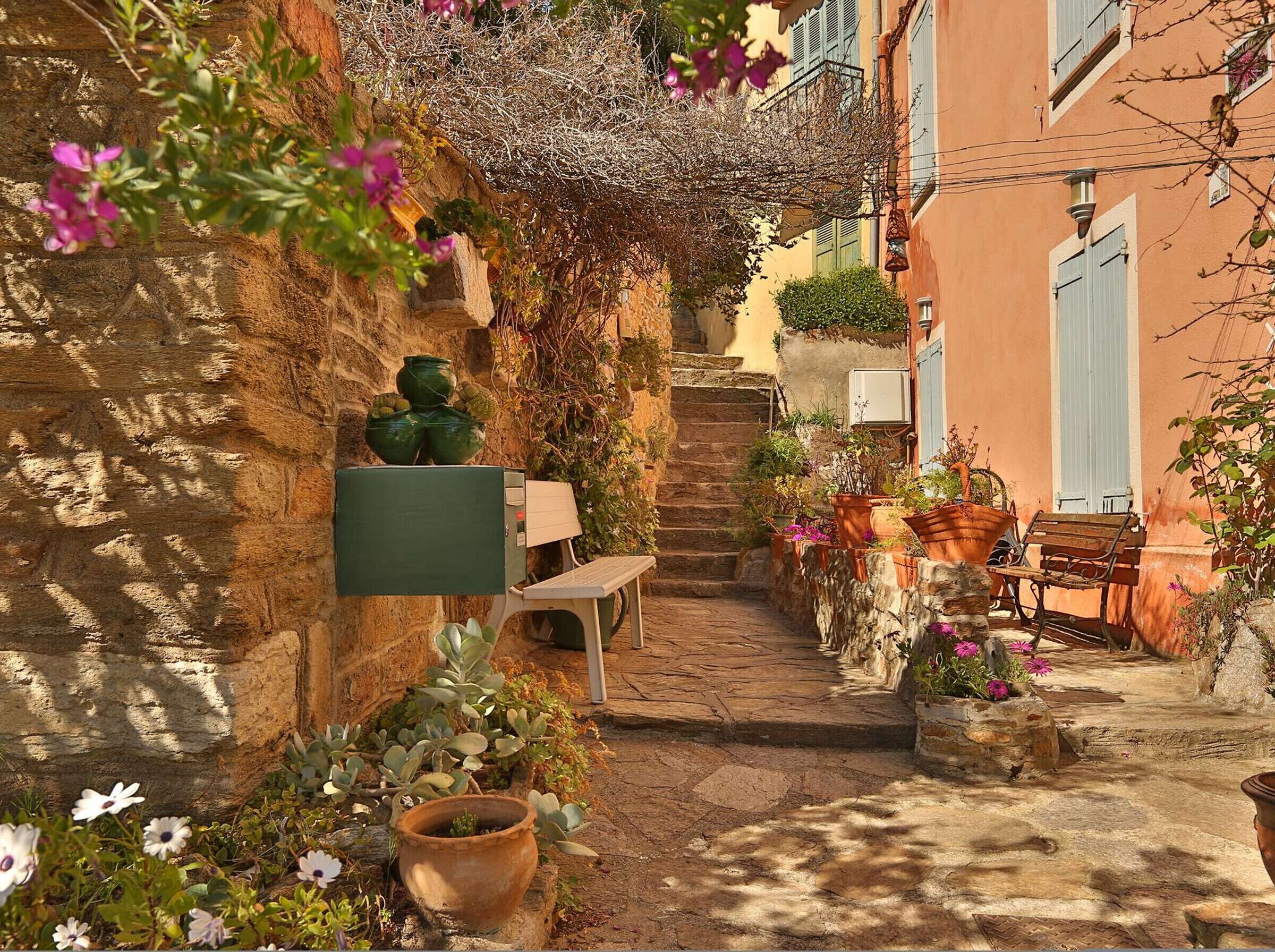 A narrow stone pathway leading to a bench and a mailbox, surrounded by potted plants and flowers.
