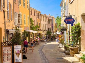 Eine enge Straße gesäumt von Geschäften und Cafés, Menschen gehen und sitzen draußen.