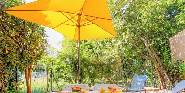 Uma área de refeições ao ar livre com um guarda-sol amarelo, mesa com cadeiras e uma cesta de pão e frutas.