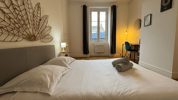 A hotel room with a large bed, a desk with a chair, a window with curtains, and a view of the city. - Apartment in Le Mans Near 24 Hours Circuit (Le Mans)