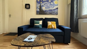 A living room with a dark blue sofa, two cushions, a wooden coffee table with a brochure, and a painting of a night sky on the wall. - Apartment in Le Mans Near 24 Hours Circuit (Le Mans)