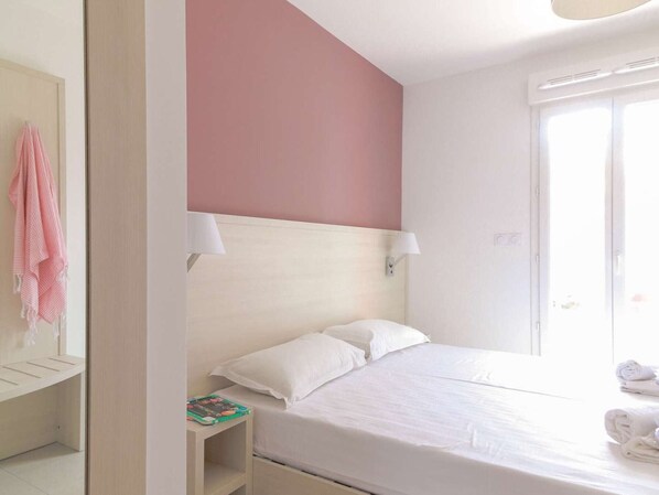 A hotel room with a bed, two bedside lamps, a nightstand with a book, a towel on the bed, and a door with a towel rack.