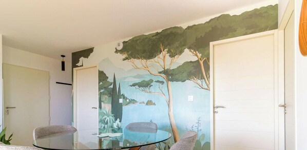 A room with a glass table, two chairs, and a mural of a landscape with trees and a body of water. - Relaxing Apartment with Terrace and Calm in Bormes-les-Mimosas (Bormes-les-Mimosas)
