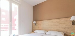 Une chambre d’hôtel avec un lit, deux lampes de chevet, une tête de lit en bois et une fenêtre donnant sur des immeubles.