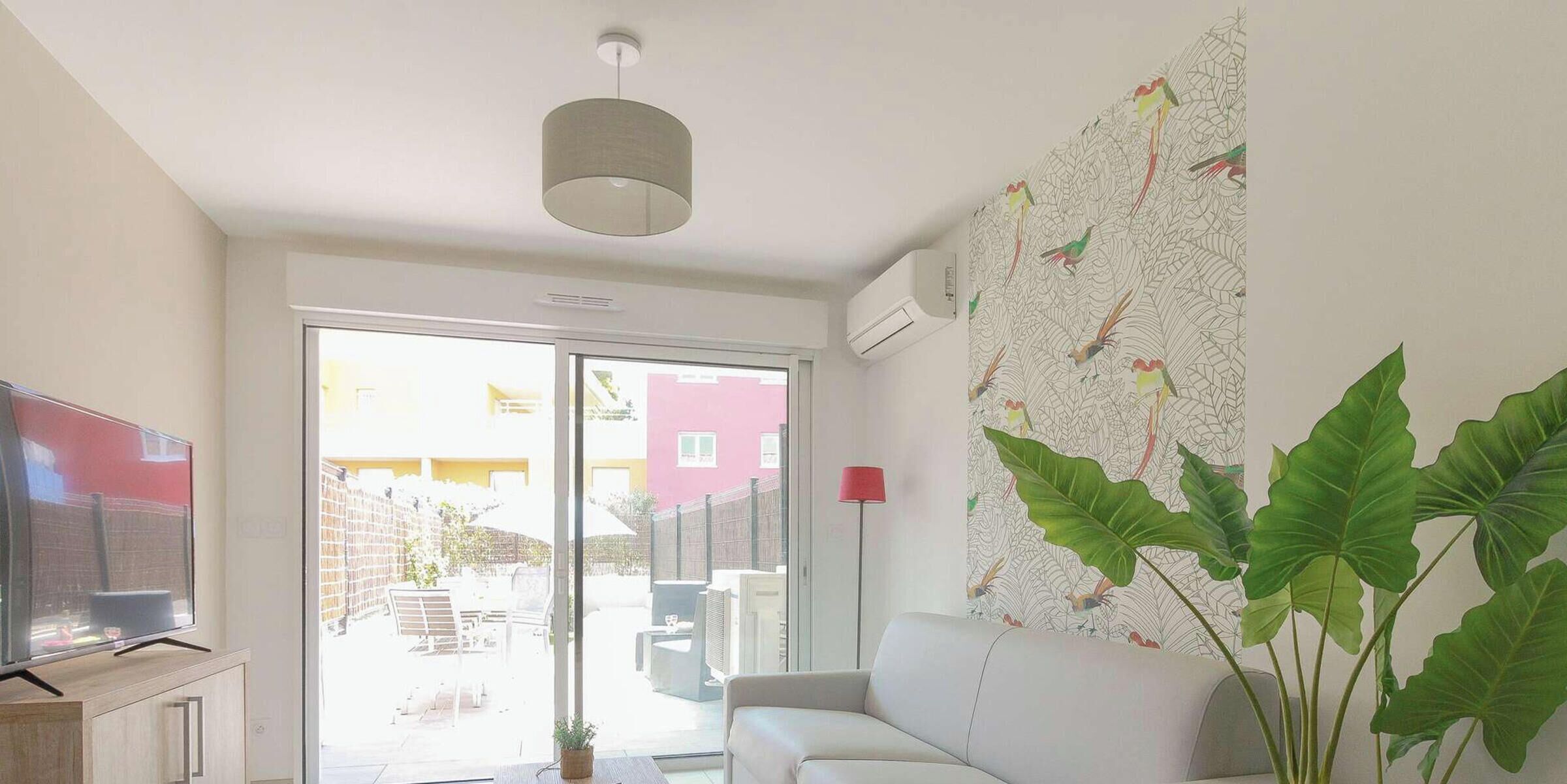 A living room with a sofa, a flat-screen TV, a ceiling light, a large window, and a wall with a leaf pattern.