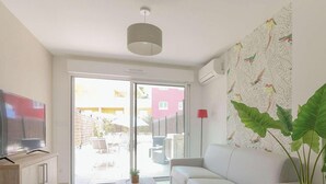 A living room with a sofa, a flat-screen TV, a ceiling light, a large window, and a wall with a leaf pattern. - Modern duplex with pool and garden, for 6 in Bormes-les-Mimosas (Bormes-les-Mimosas)