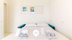 A hotel room with a bed, two pillows, a blue headboard, and a pair of slippers on the bed. There are three framed pictures on the wall above the bed.