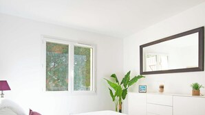 A bedroom with a large window, a bed, a dresser, a mirror, and a potted plant.