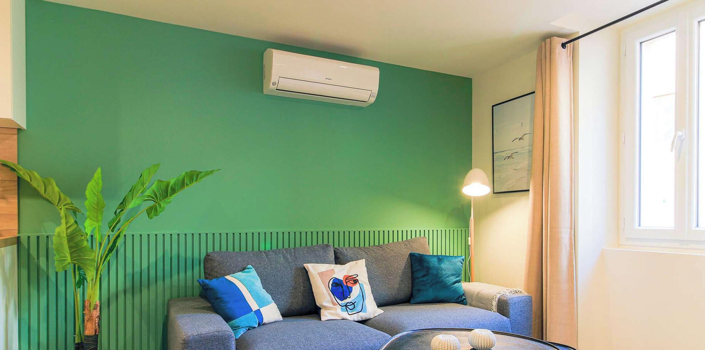 Ein Zimmer mit grüner Wand, einem grauen Sofa mit Kissen, einer Topfpflanze und einem Fenster mit Vorhängen.