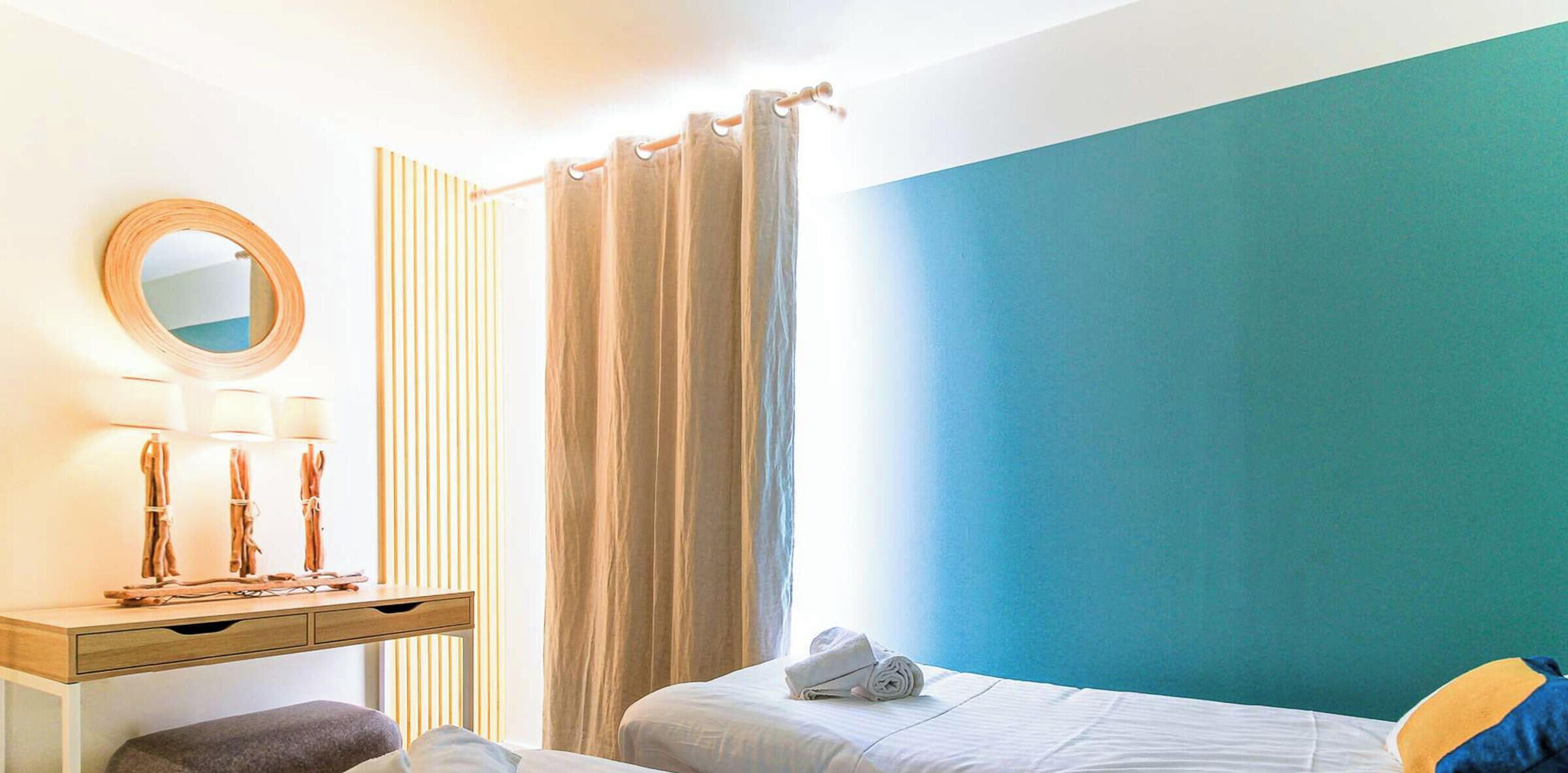 Quarto de hotel com cama, escrivaninha com espelho e janela com cortinas.