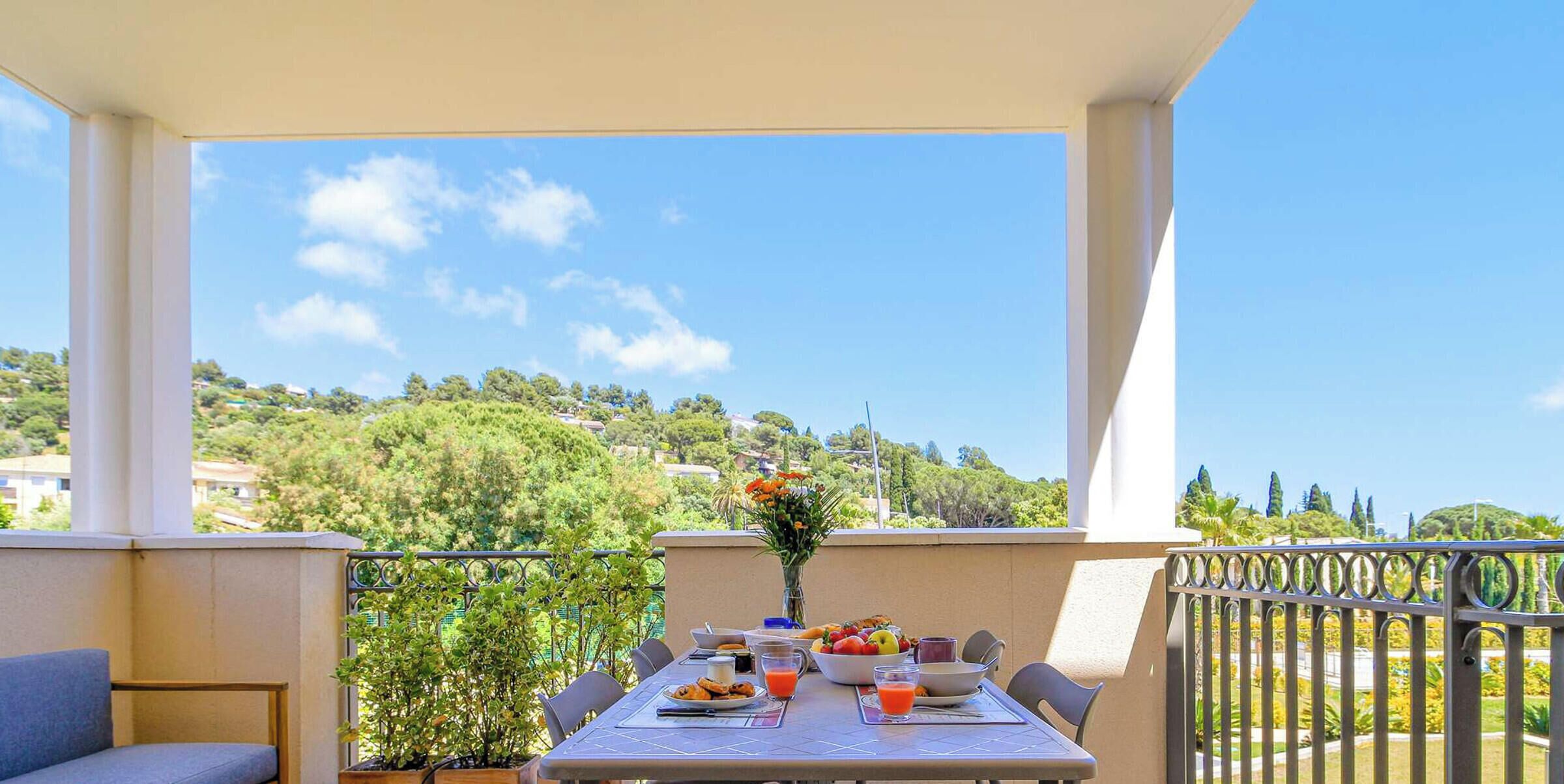 Un balcón con una mesa lista para el desayuno, una planta en maceta y vistas a una ladera.
