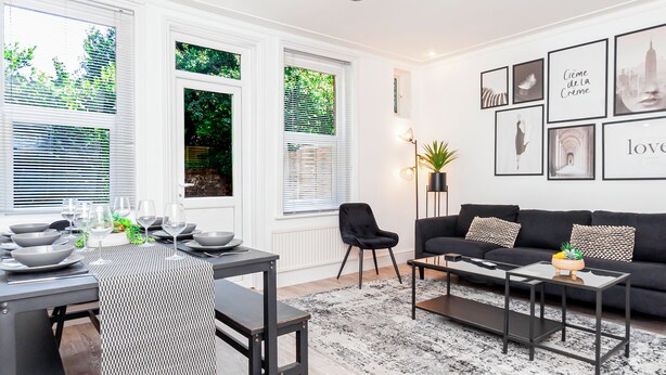Een moderne woonkamer met een eethoek, met een zwarte bank, een salontafel, een eettafel met kommen en wijnglazen, en kunstwerken in lijsten aan de muur.