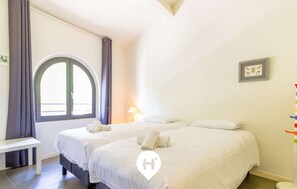 A hotel room with two beds, a bench, a window with curtains, a lamp, and a framed picture on the wall.