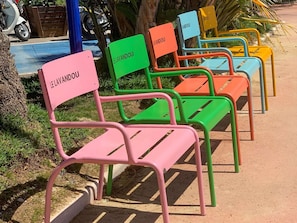 A row of colorful metal chairs with "LE LAVANDOU" signs, arranged on a concrete path with greenery and a parked scooter in the background.