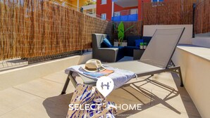 A sunlit outdoor seating area with a lounge chair, a table with a hat and sunglasses, and a bamboo fence.