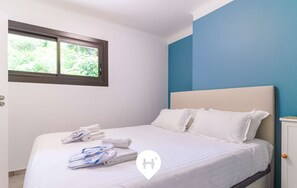 A neatly made bed with white linens and pillows, a window with a view of greenery, and a blue accent wall.