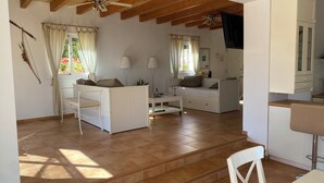 A spacious living area with a high ceiling, wooden beams, a white sofa, a dining table, and a kitchen area with cabinets and a TV. - Charming 2-bedroom villa in Santa Margalida with AC, WiFi, fitness room (Santa Margalida)