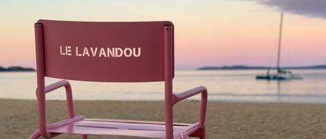 Ein rosafarbener Stuhl mit dem Aufdruck "LE LAVANDOU" an der Rückenlehne, platziert an einem Sandstrand mit ruhiger See und einem Segelboot im Hintergrund.