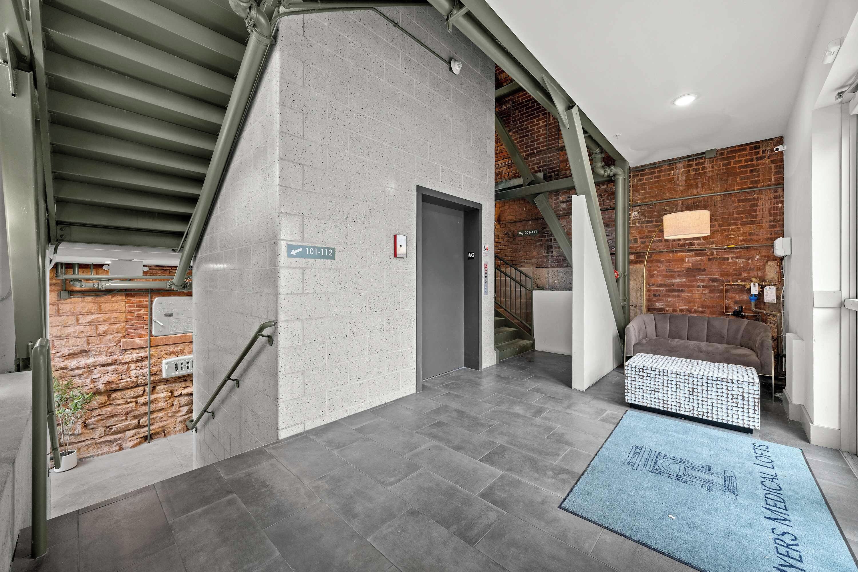 A modern lobby with a staircase, a grey tiled floor, a blue medical logo mat, and a brick wall.