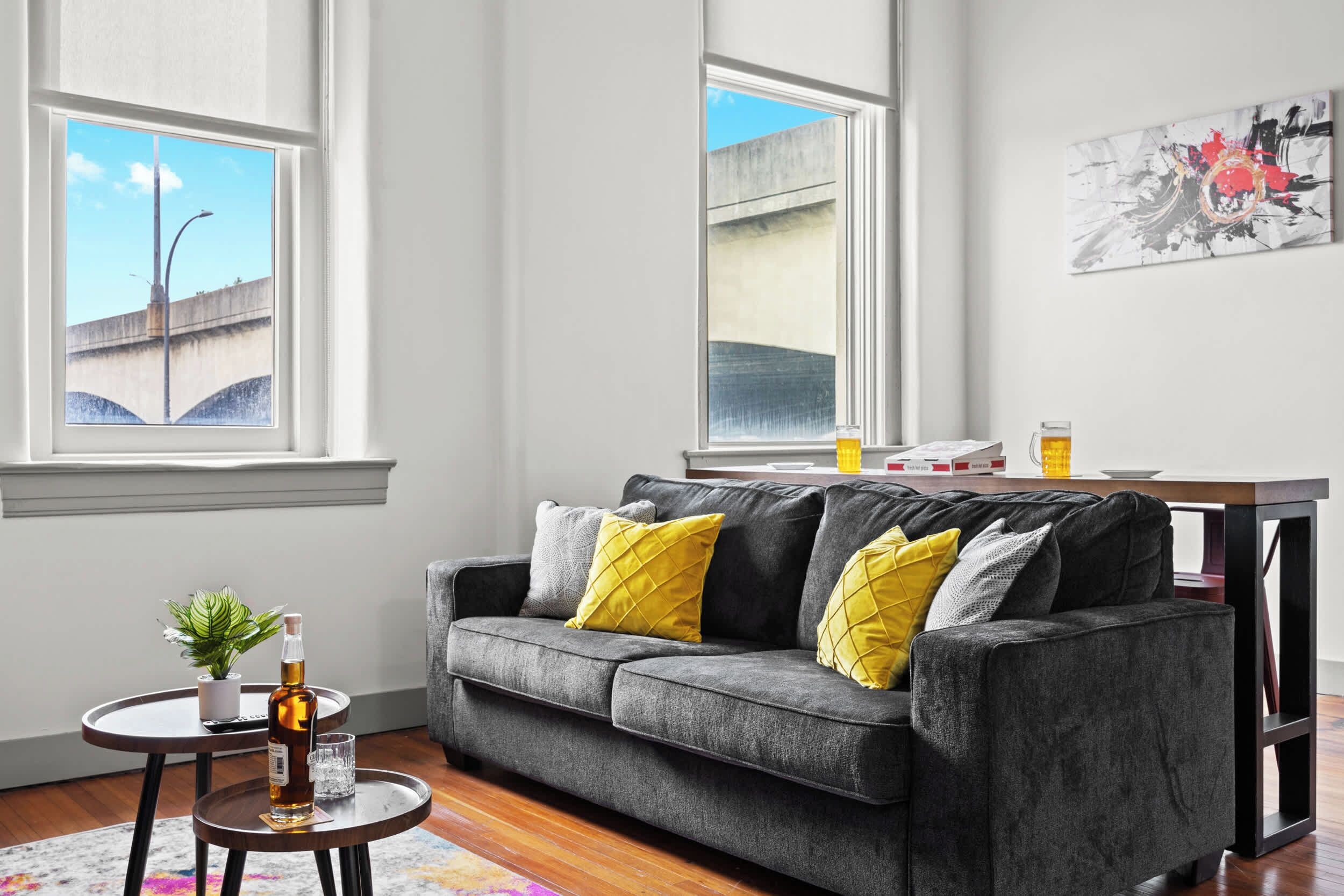 A modern living room with a grey sofa, yellow cushions, a small round table with a bottle and glass, and a painting on the wall.