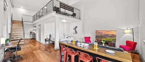 Un vaste espace de vie doté d’un plancher en bois, d’un bar central avec des tabourets rouges, d’un escalier menant à un balcon et d’un grand tableau représentant un stade.