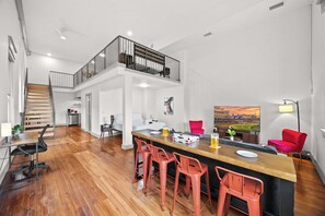 Un vaste espace de vie doté d’un plancher en bois, d’un bar central avec des tabourets rouges, d’un escalier menant à un balcon et d’un grand tableau représentant un stade.