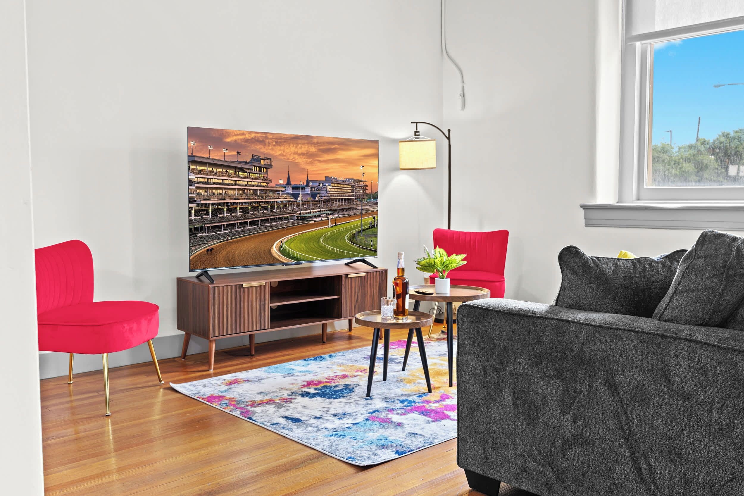 A living room with a large painting of a stadium, a red chair, a grey sofa, a small round table with a lamp, and a wooden cabinet.