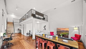 A spacious living area with a wooden floor, a central bar with red bar stools, a staircase leading to a balcony, and a large painting of a stadium. - Chic & comfy unit in Louisville's Medical District (Louisville)