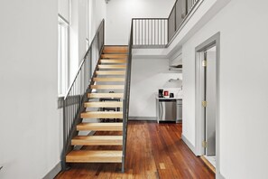 Un escalier avec des marches en bois et des rampes en métal mène à un espace cuisine doté de meubles blancs et d’un appareil en acier inoxydable.