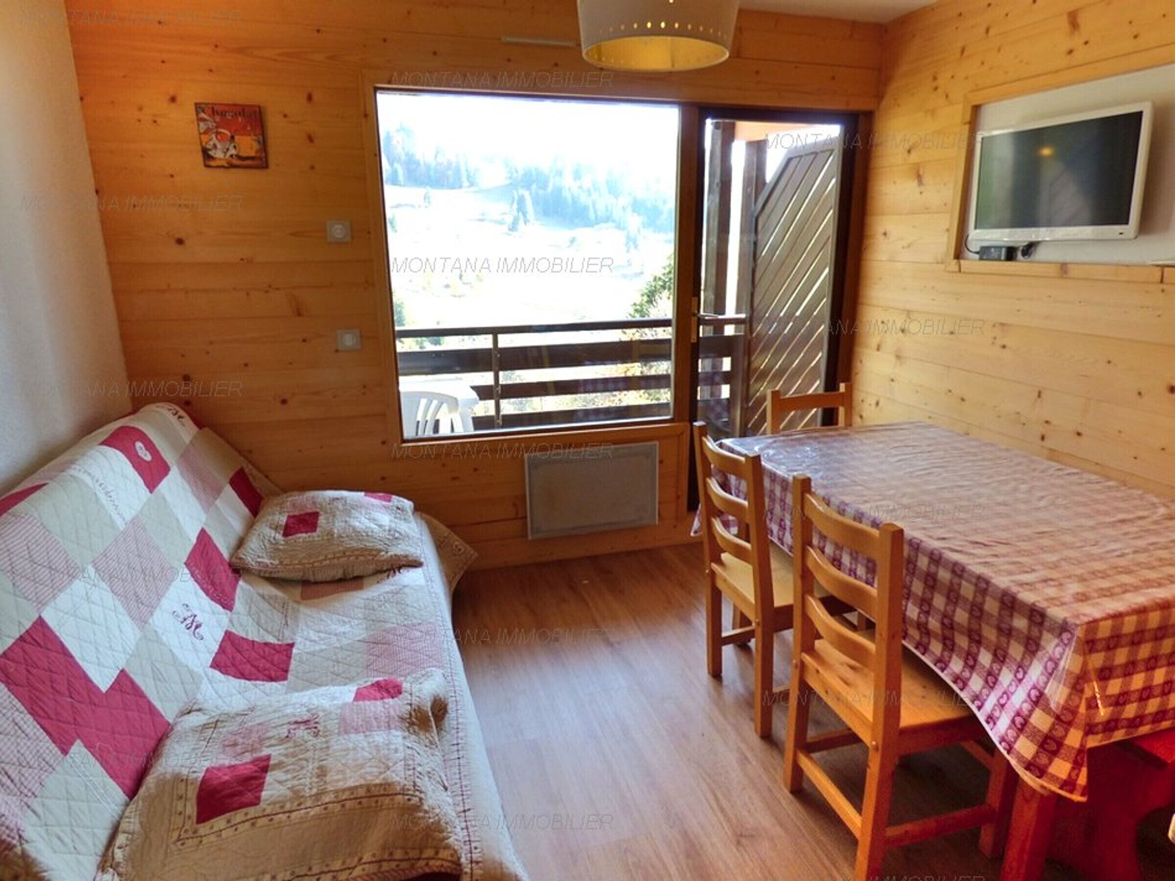 A cozy cabin interior with a wooden floor, a quilted sofa, a table with a checkered tablecloth, and a TV mounted on the wall.