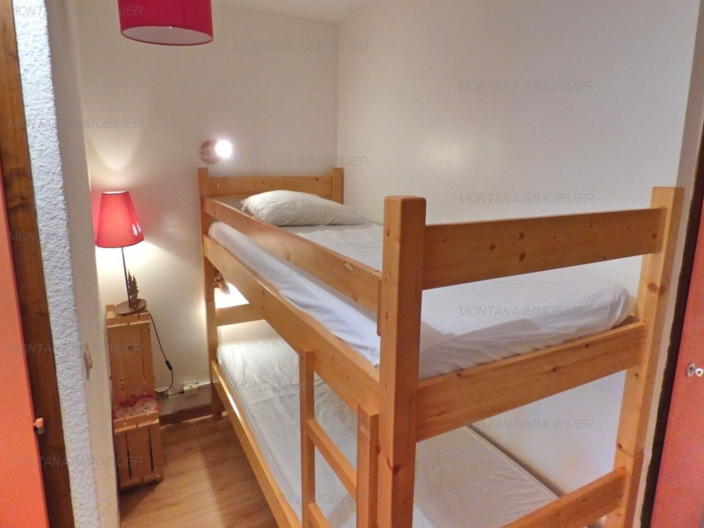 Une chambre avec un lit superposé doté d’une tête de lit en bois, une table de chevet avec une lampe et un mur blanc orné d’un motif discret.